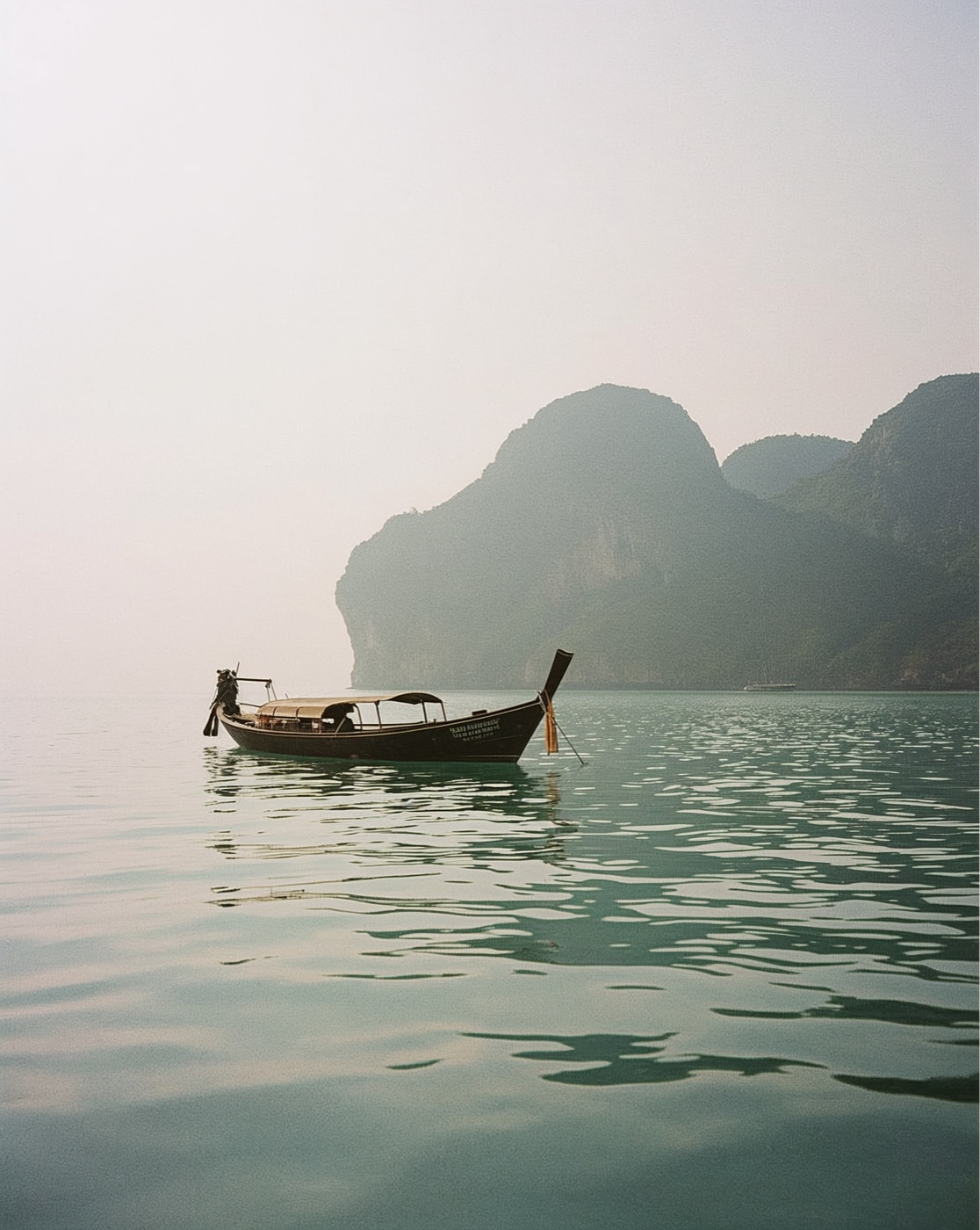 A wooden longtail boat on still water below limestone cliffs in the morning haze
